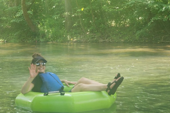 Person relaxing on a green float in a river, surrounded by trees, waving and smiling.