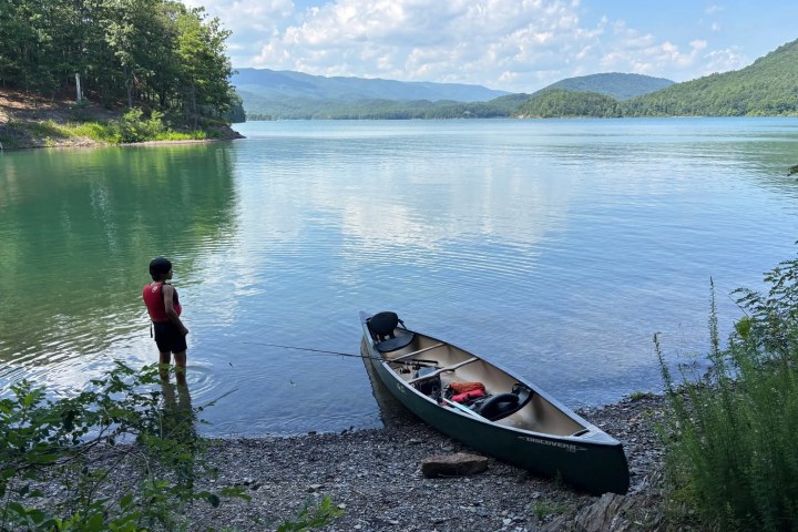 Person in life vest stands near a canoe on a lake shore with distant mountains.