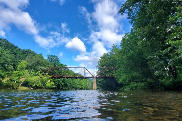 A rusty metal bridge spans over a tree-lined river under a blue sky with scattered clouds.