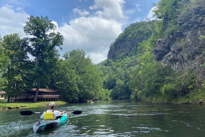Person kayaking on a river with trees and a rocky cliff in the background under a partly cloudy sky.