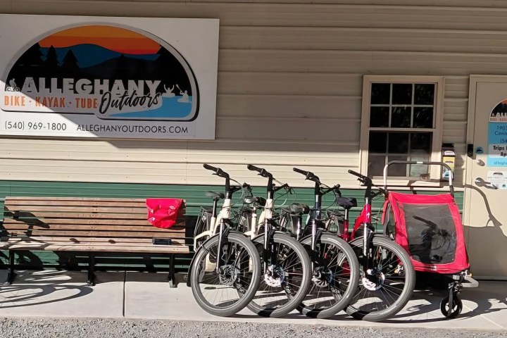 Bicycles and a red trailer parked near a bench and Alleghany Outdoors sign on a building.