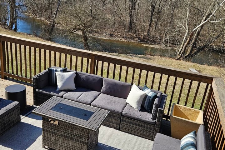 Patio with wicker furniture set and fire pit table overlooking a river with bare trees.